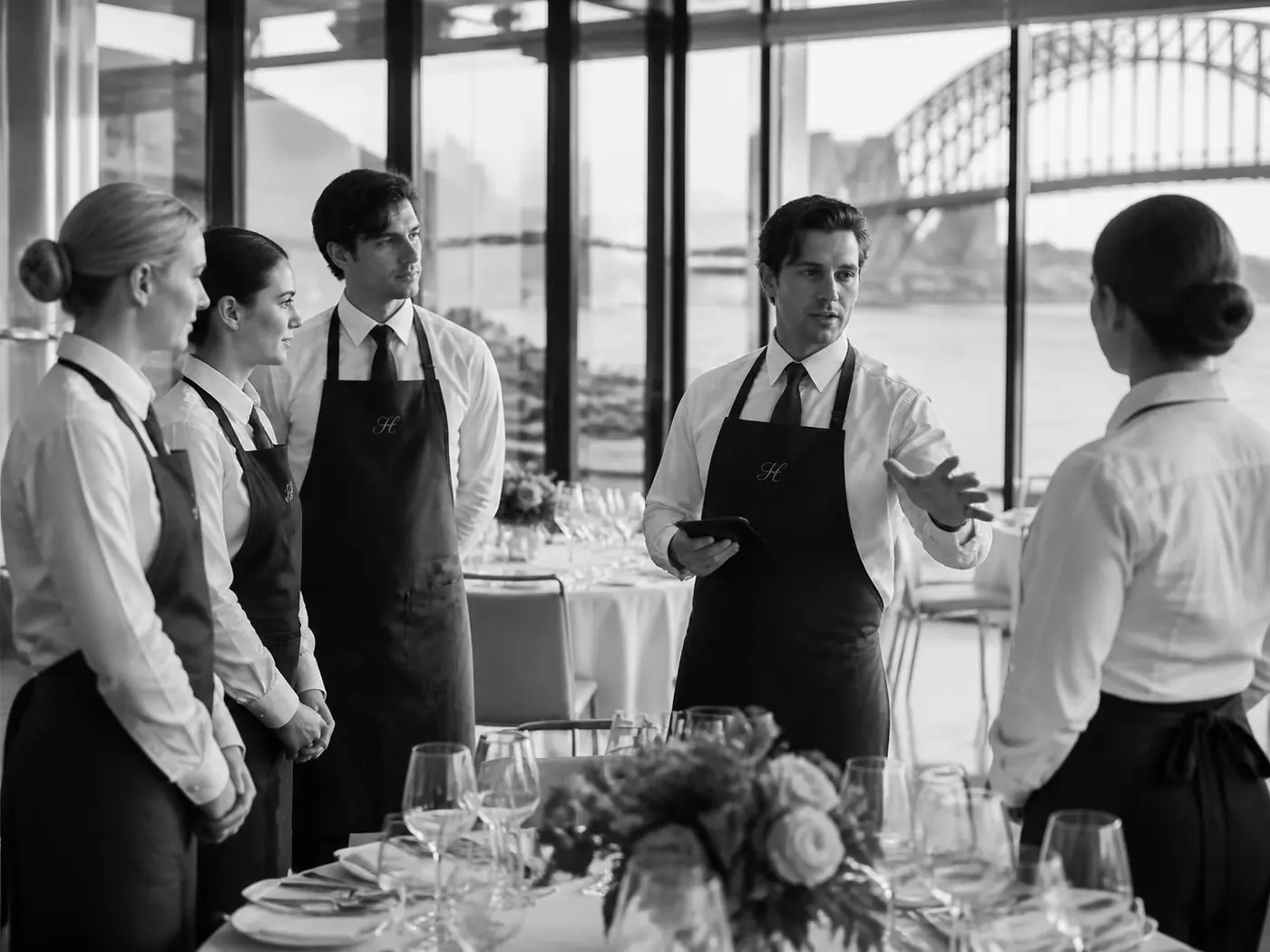Opera View event team in white shirts and deep navy monogrammed aprons briefing before a harbourfront event, in black and white